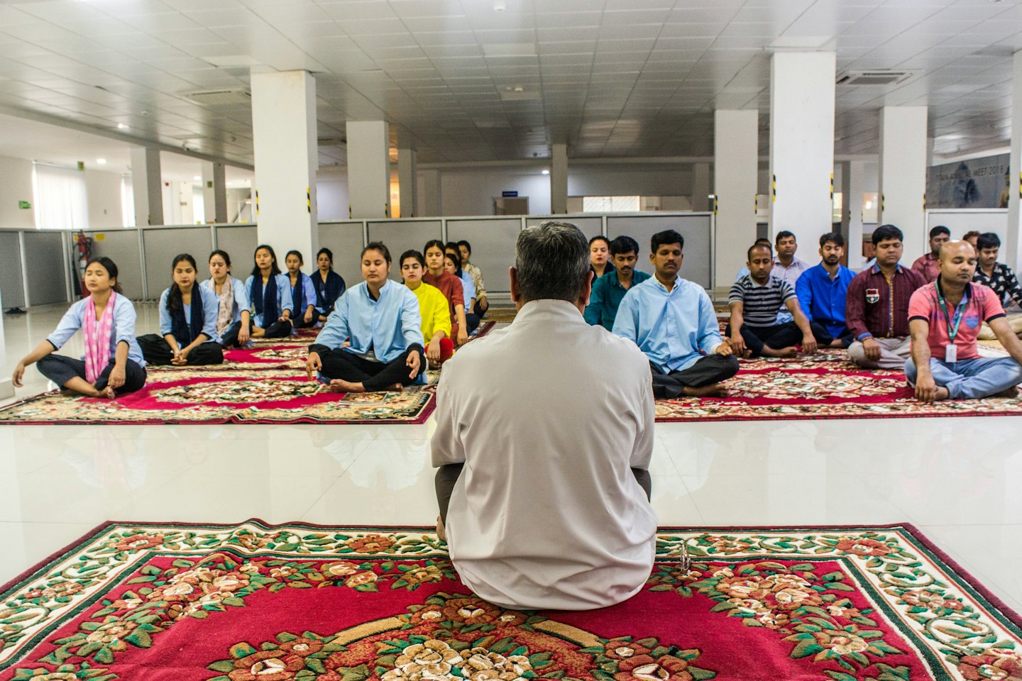 Persone in meditazione a gambe incrociate su tappeti rossi in un ufficio luminoso.